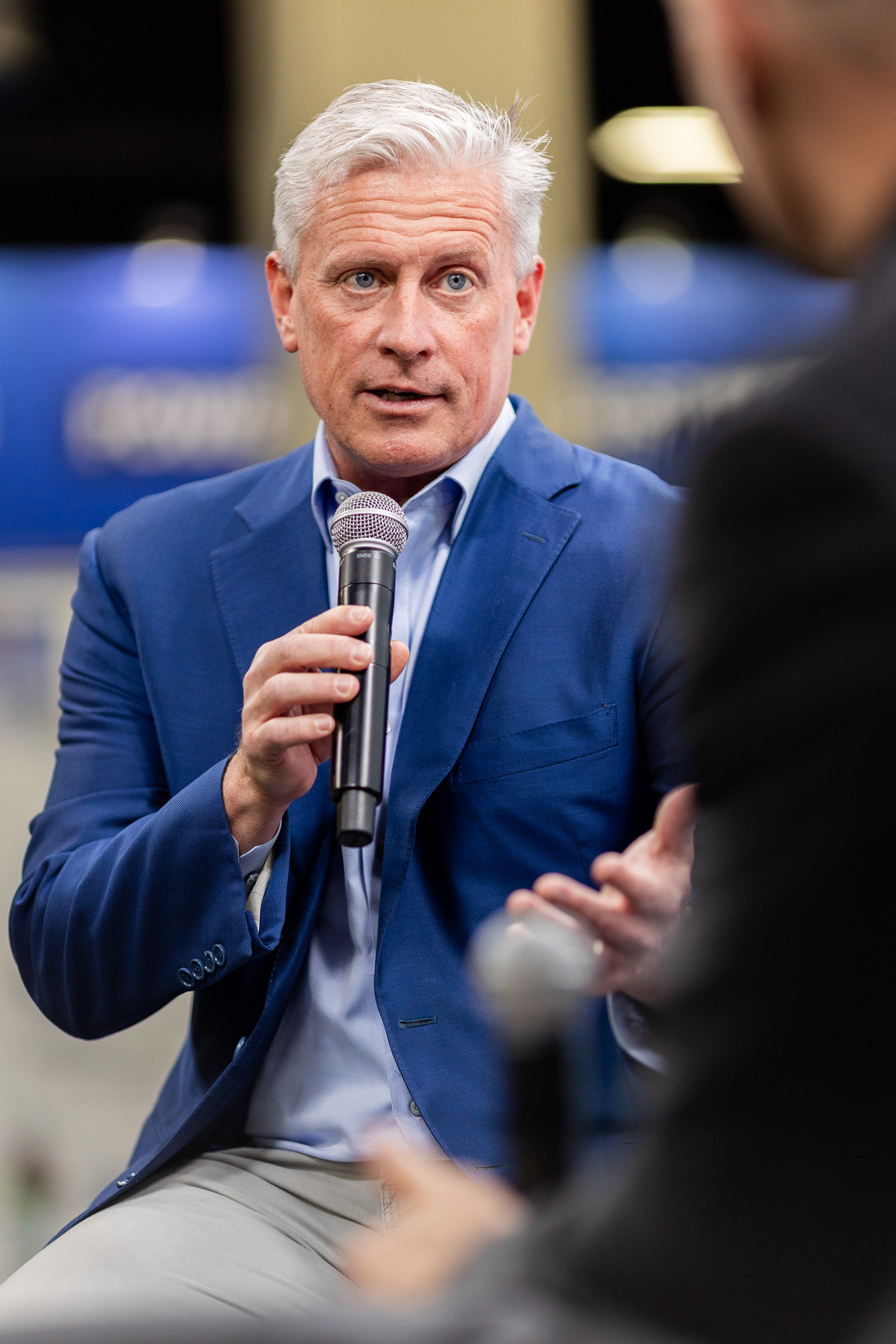 A man with gray hair in a blue suit jacket speaks into a microphone during an interview or discussion. Another person, blurred in the foreground, also holds a microphone. The background is indoors and slightly out of focus.