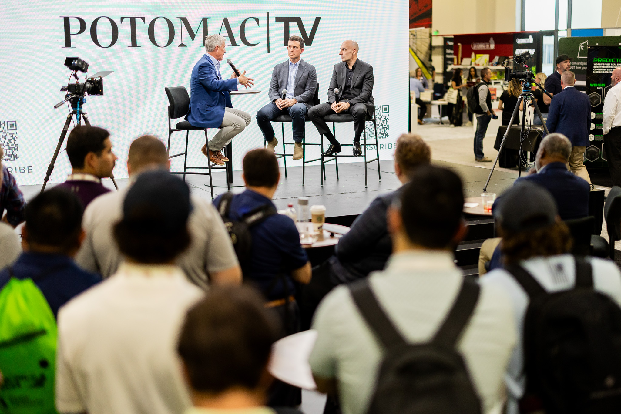 A group of people watch as three men sit on stage having a discussion in front of a large screen that reads "POTOMAC TV." Cameras and recording equipment are set up around the stage.