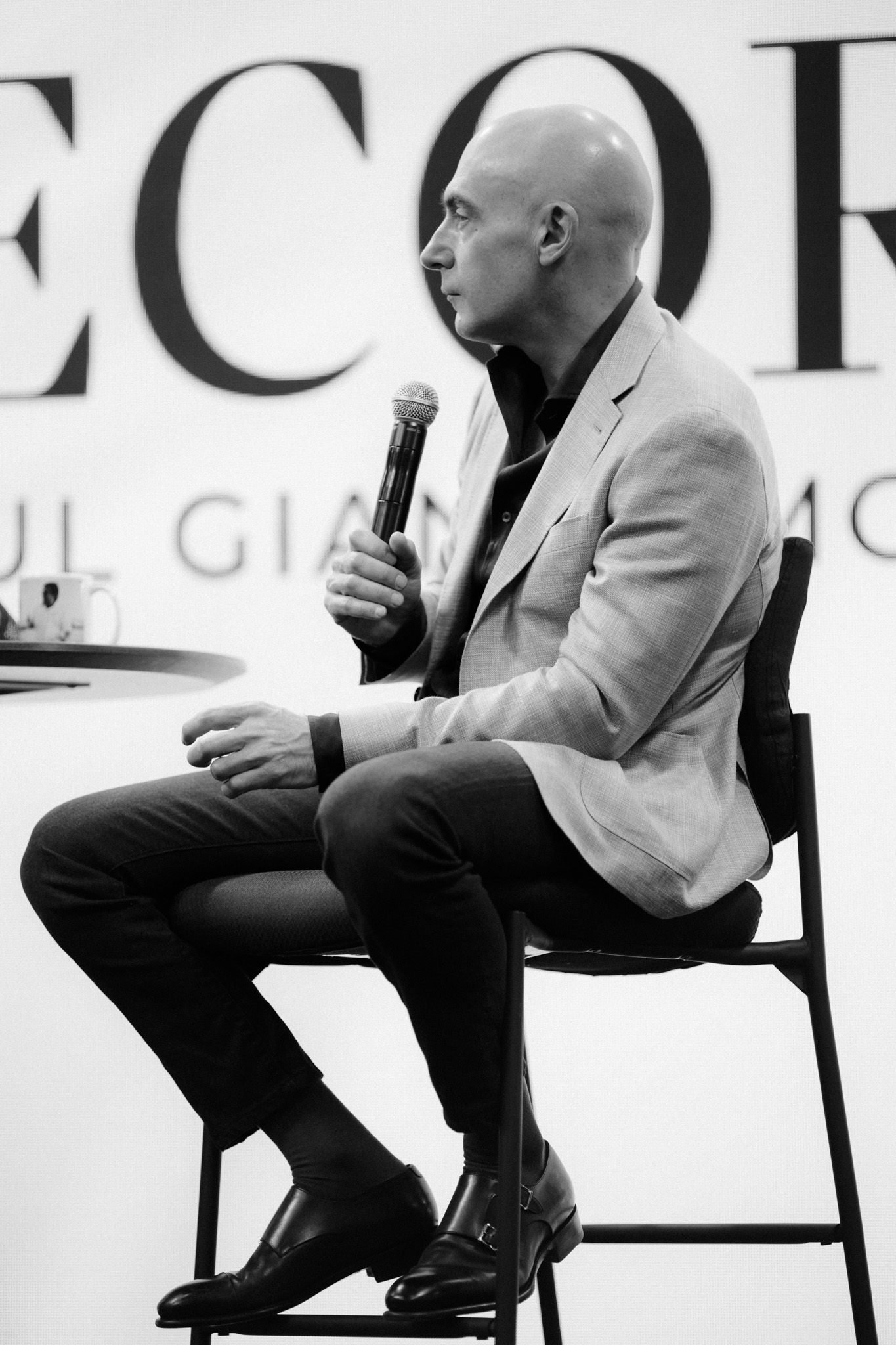 A bald man in a blazer and dress shoes sits on a high chair, holding a microphone and speaking, captured in black and white. The background shows large, partially visible text.
