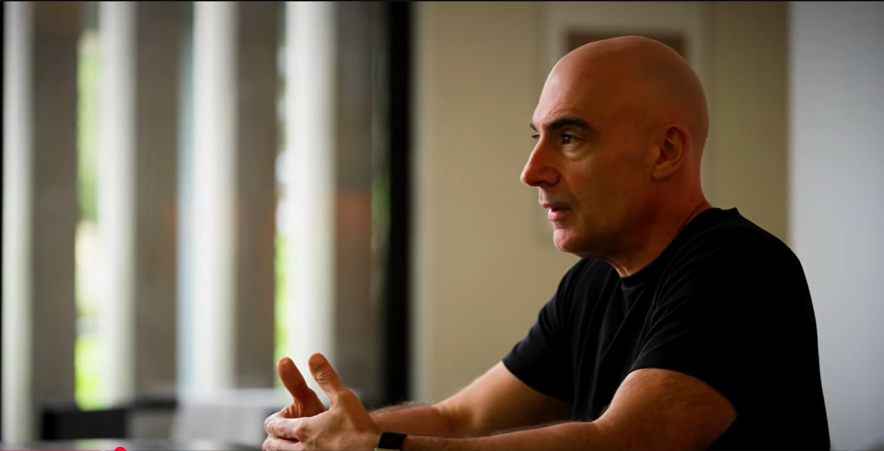 A bald man in a black t-shirt sits indoors at a table, gesturing with his hands while speaking. Bright windows with vertical blinds are in the background, creating a softly lit atmosphere.