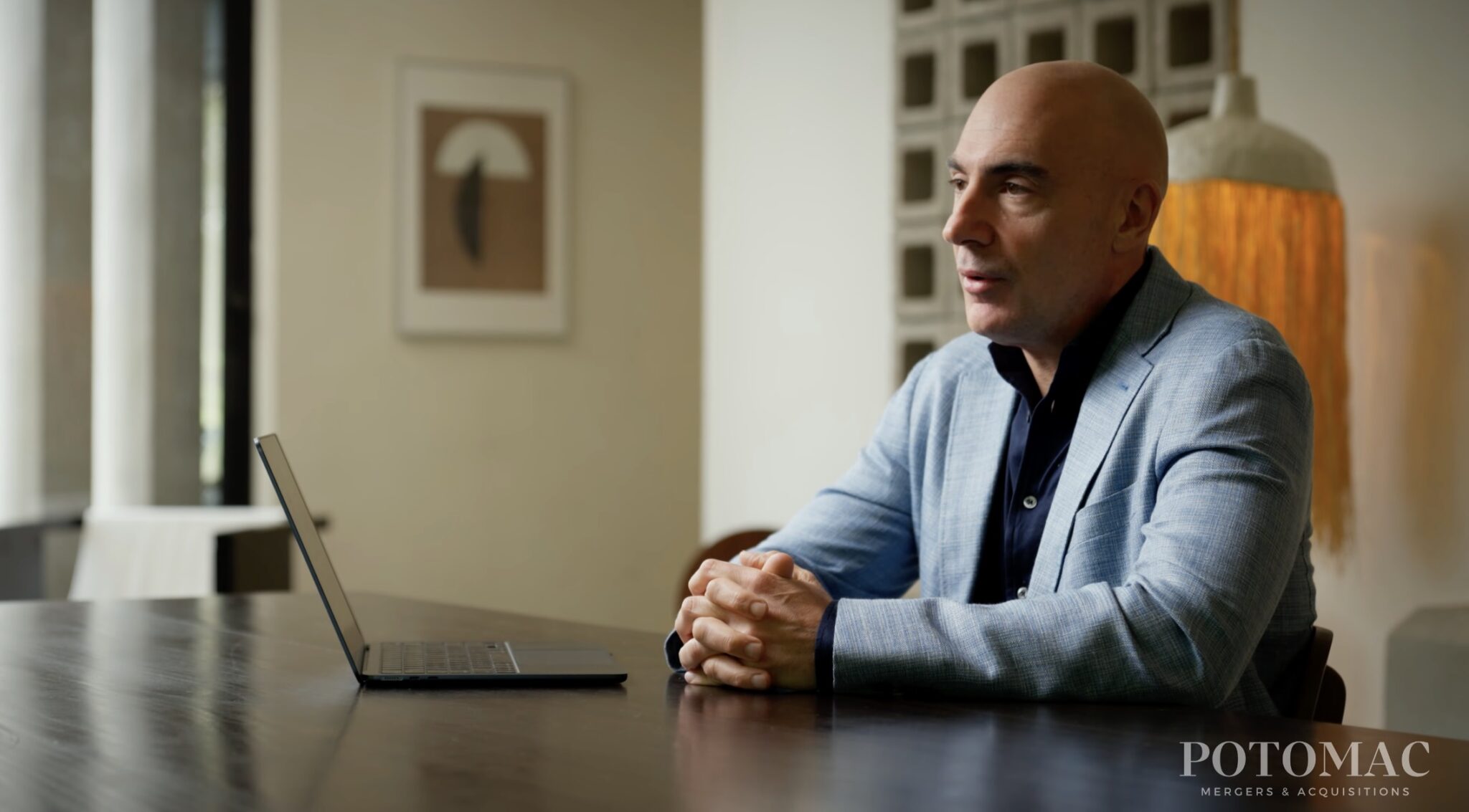 A man in a blue blazer sits at a table with a laptop, hands clasped, speaking. The background features modern decor. The logo "POTOMAC MERGERS & ACQUISITIONS" appears in the bottom right corner.
