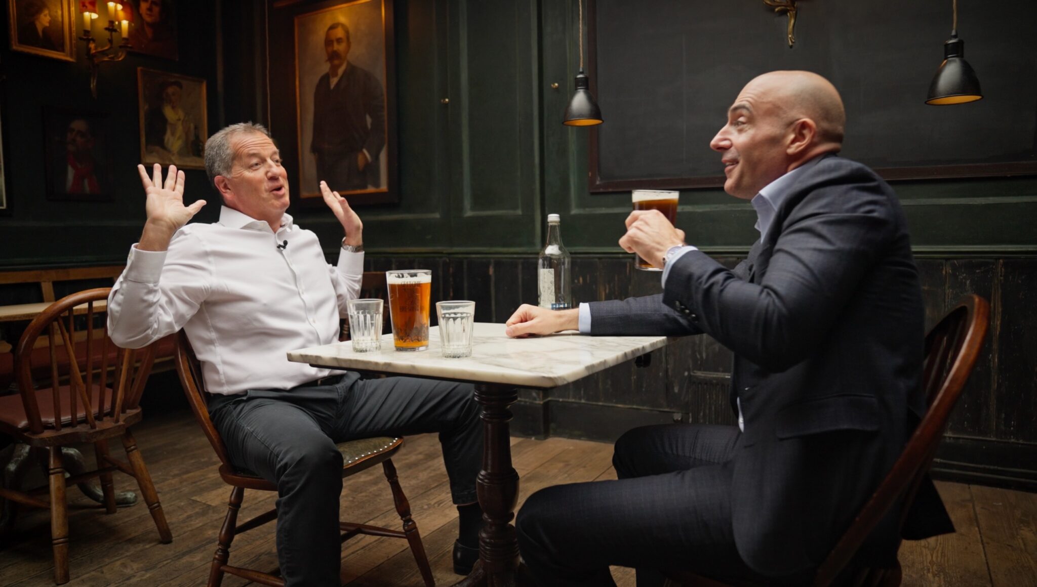 Two men sit at a small table in a pub, each with a pint of beer. One man gestures animatedly with raised hands, while the other smiles, holding his drink. Framed portraits hang on the dark green walls behind them.