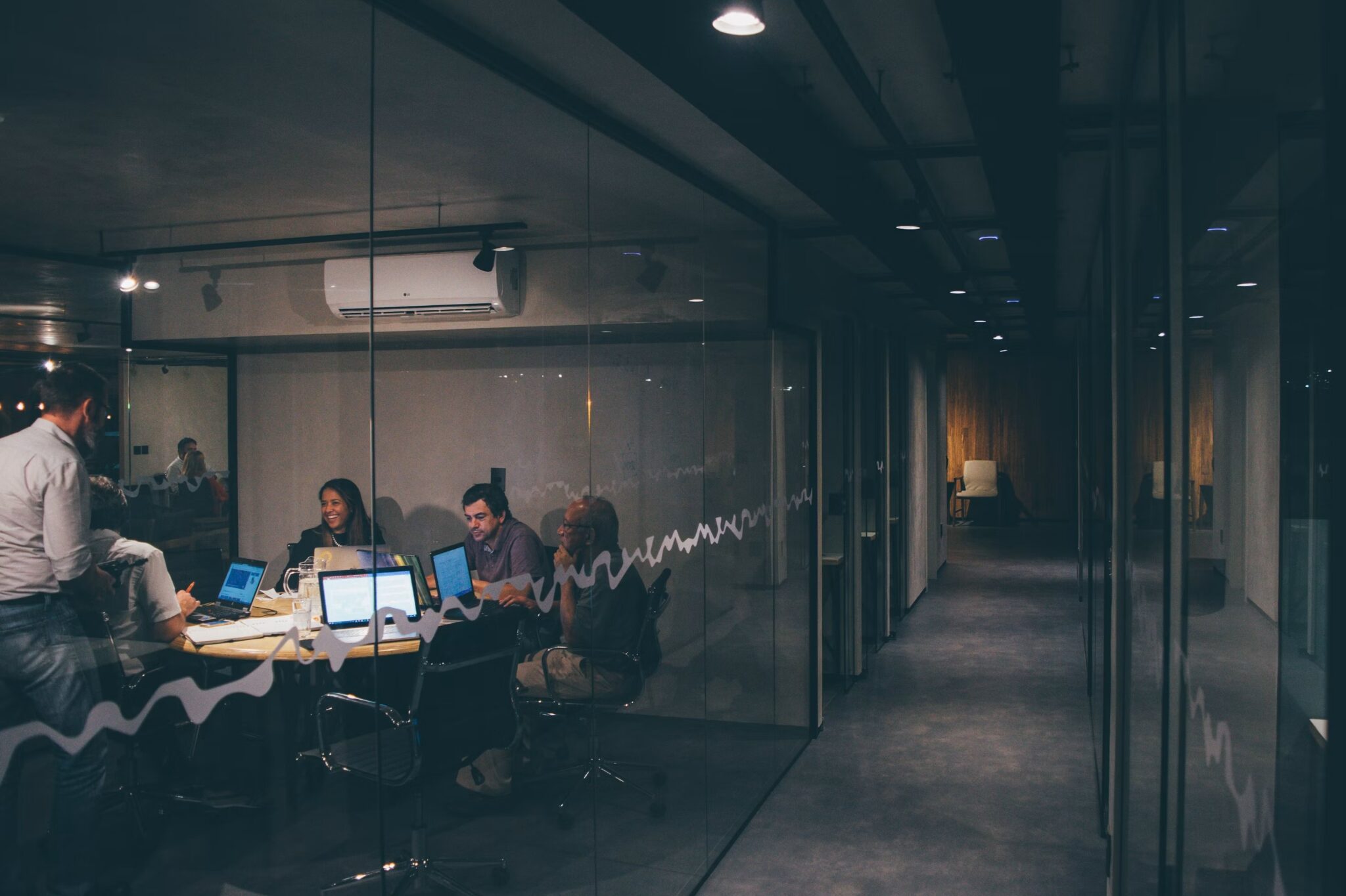 A group of people sit around a conference table with laptops in a glass-walled office. The room is dimly lit, and there is a long empty hallway visible through the glass.