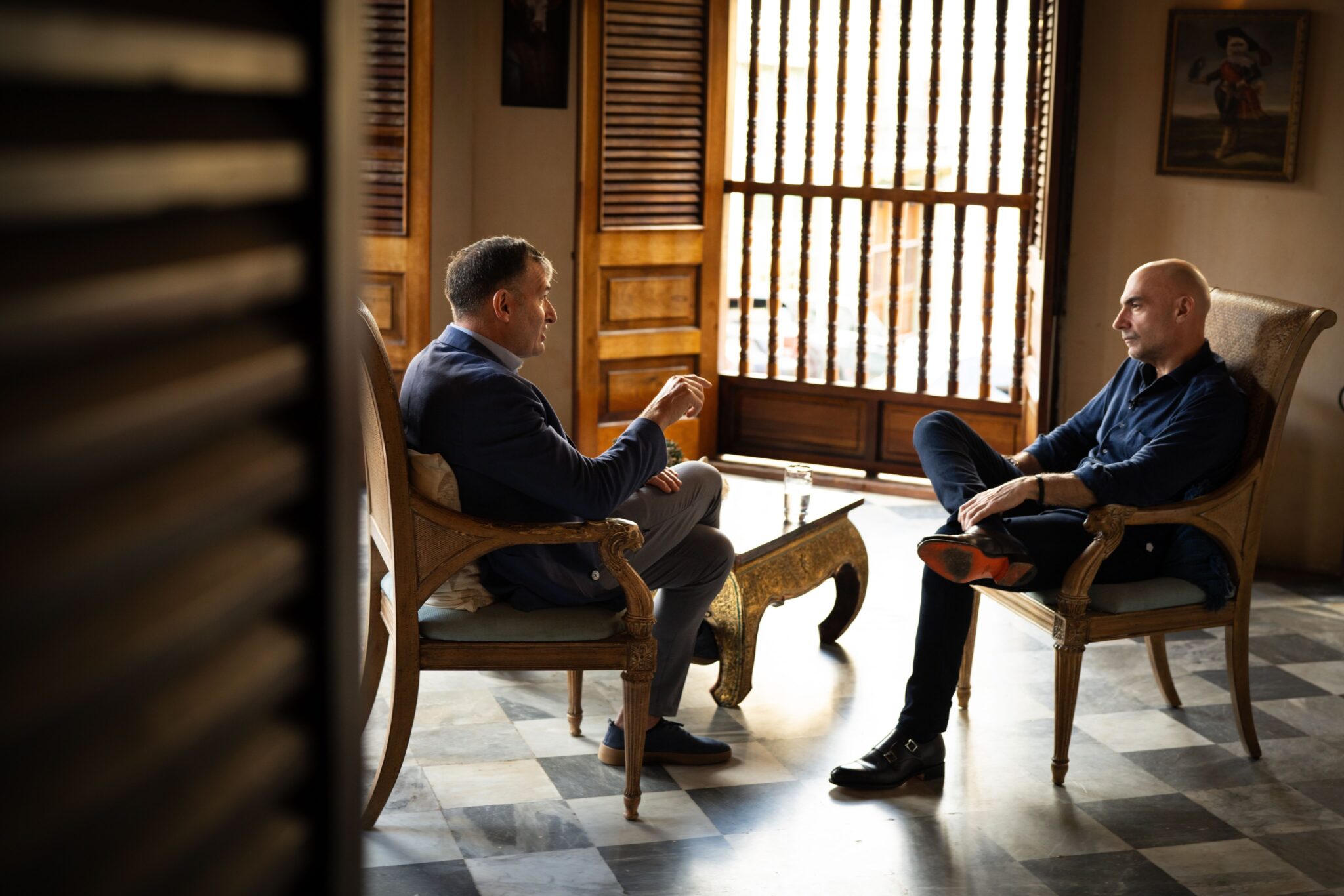 Two men sit facing each other in wooden chairs, engaged in conversation in a sunlit room with wooden shutters and marble floors. One man gestures with his hand while the other listens attentively with legs crossed.
